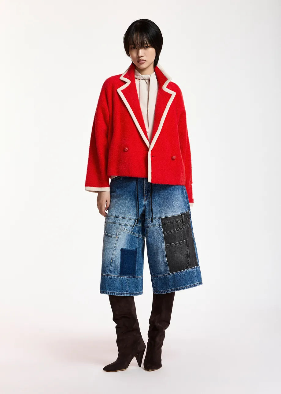 A model stands against a plain white studio background wearing a bright red cropped double-breasted blazer with white piping, blue patchwork denim culottes, and dark brown knee-high boots.