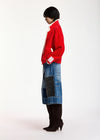 A model shown from the side profile wearing a bright red knit cardigan with white trim, a blue patchwork denim skirt, and dark brown knee-high heeled boots, set against a plain white studio background.