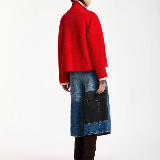 A model shown from the back wearing a bright red cropped cardigan, a blue denim patchwork skirt with a black pocket, and dark brown boots, against a plain white studio background.
