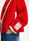 A close-up of a person wearing a bright red fuzzy jacket with cream-colored trim and a visible patch on the sleeve. The person is also wearing blue jeans, set against a plain white background.