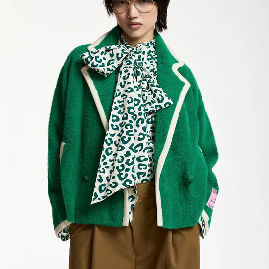 A model wearing a green fuzzy cropped cardigan with cream piping, a green and white leopard-print bow tie-neck blouse, and brown trousers, photographed against a plain white studio background.