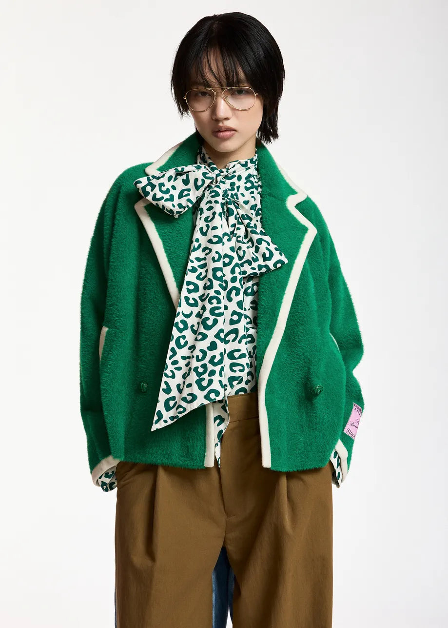 A model wearing a green fuzzy cropped cardigan with cream piping, a green and white leopard-print bow tie-neck blouse, and brown trousers, photographed against a plain white studio background.