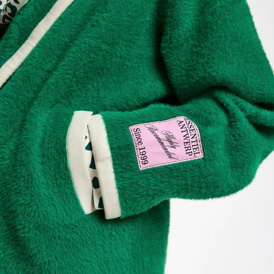Close-up of a green fuzzy cardigan with cream ribbed trim, a pink brand patch on the sleeve, and a patterned shirt underneath, paired with blue jeans.