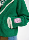 Close-up of a green fuzzy cardigan with cream ribbed trim, a pink brand patch on the sleeve, and a patterned shirt underneath, paired with blue jeans.