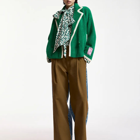 A model stands against a plain light background wearing a bright green cropped jacket with white piping, a green and white patterned scarf, wide-leg brown trousers with blue side panels, and red shoes.