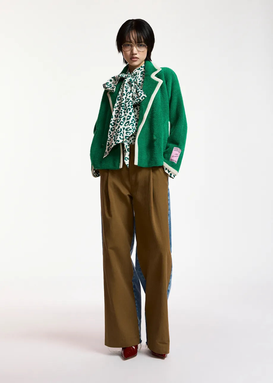 A model stands against a plain light background wearing a bright green cropped jacket with white piping, a green and white patterned scarf, wide-leg brown trousers with blue side panels, and red shoes.