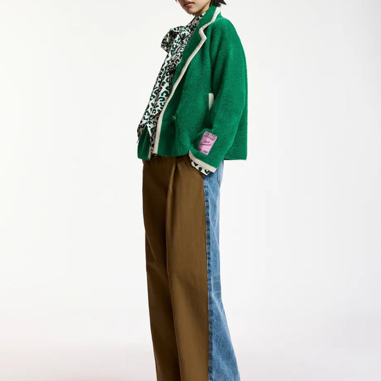 A model stands in profile wearing a green knit cardigan with white trim, a patterned scarf, wide-leg brown pants, and blue denim jeans with a color-blocked side seam, against a plain white studio background.