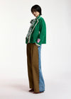 A model stands in profile wearing a green knit cardigan with white trim, a patterned scarf, wide-leg brown pants, and blue denim jeans with a color-blocked side seam, against a plain white studio background.