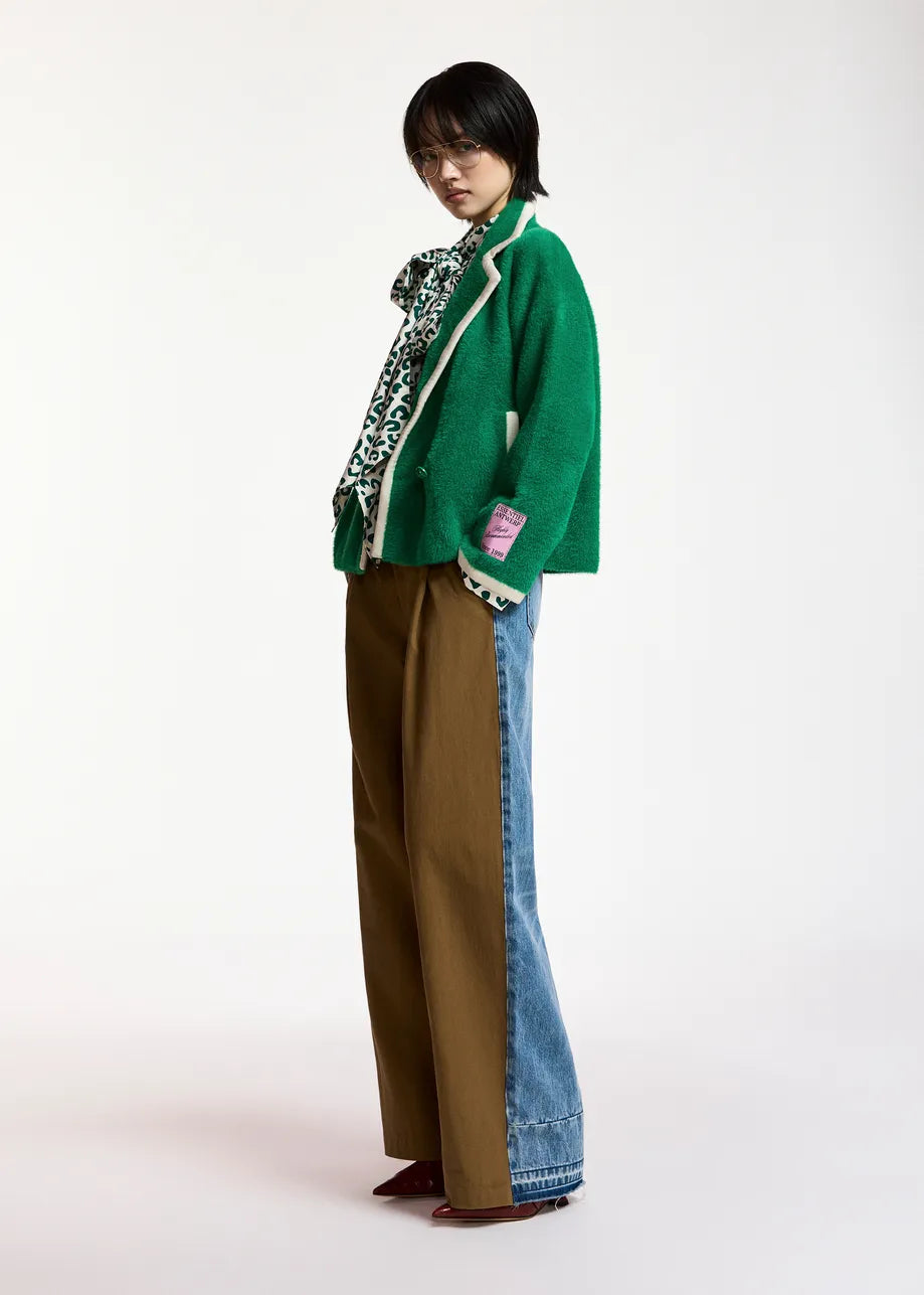 A model stands in profile wearing a green knit cardigan with white trim, a patterned scarf, wide-leg brown pants, and blue denim jeans with a color-blocked side seam, against a plain white studio background.