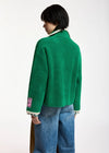 A model shown from the back wearing a bright green fuzzy knit cardigan with white ribbed trim and a pink patch on the sleeve, paired with blue jeans against a plain white background.
