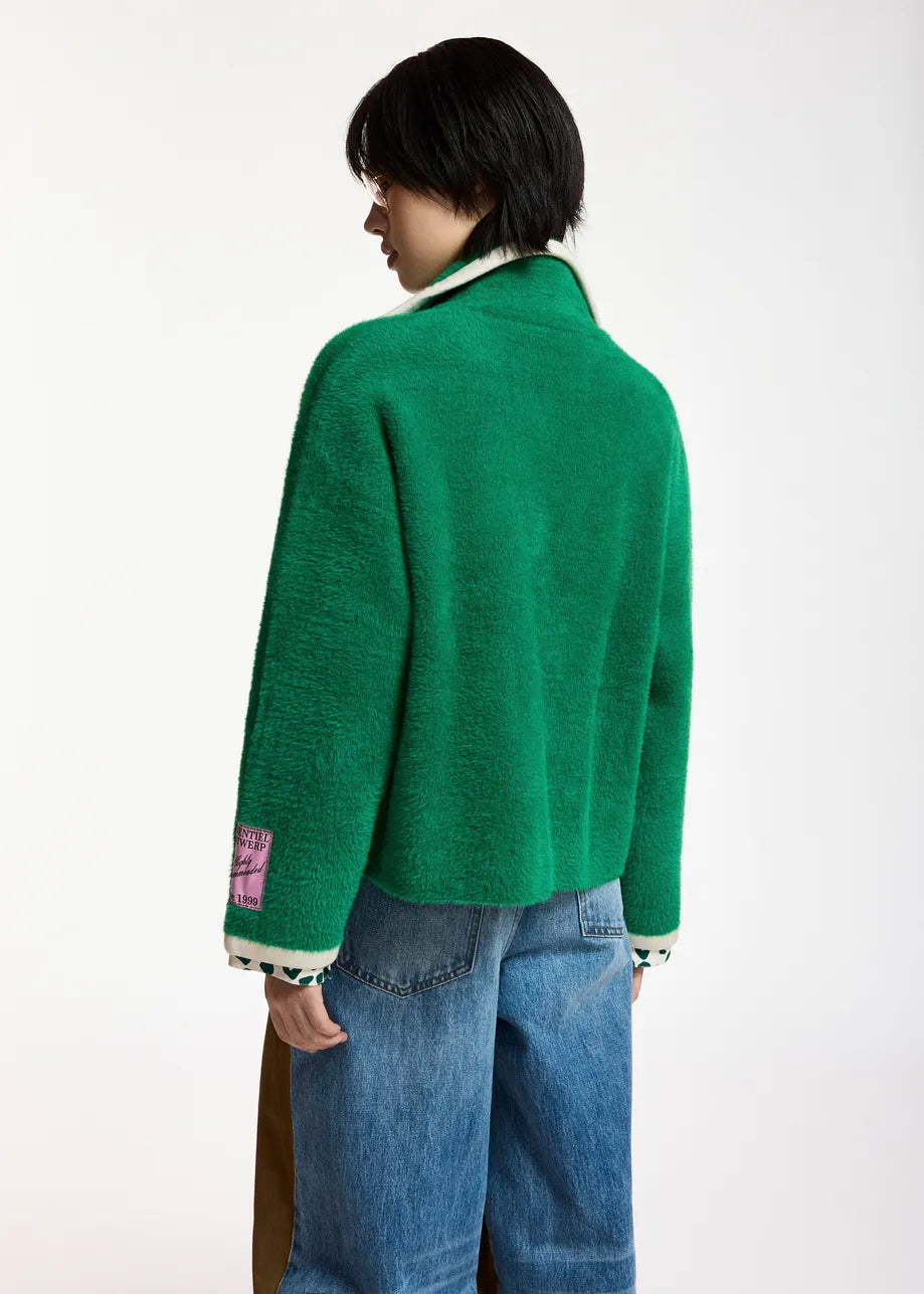 A model shown from the back wearing a bright green fuzzy knit cardigan with white ribbed trim and a pink patch on the sleeve, paired with blue jeans against a plain white background.