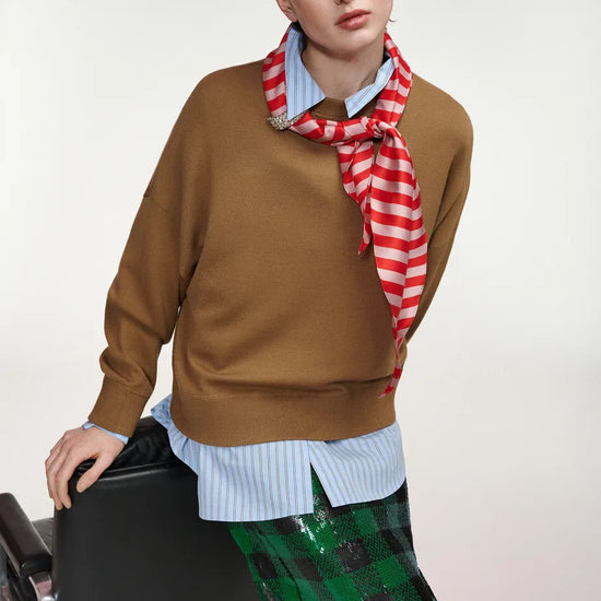 A model wearing a brown knit sweater over a light blue striped shirt, accessorized with a red and white striped scarf, and paired with a green sequined plaid skirt, leaning against a black chair against a light gray background.