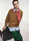 A model wearing a brown knit sweater over a light blue striped shirt, accessorized with a red and white striped scarf, and paired with a green sequined plaid skirt, leaning against a black chair against a light gray background.
