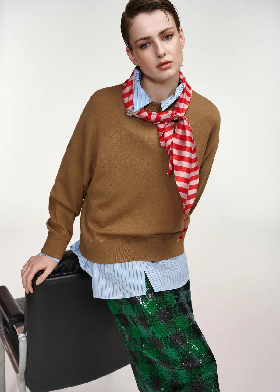 A model wearing a brown knit sweater over a light blue striped shirt, accessorized with a red and white striped scarf, and paired with a green sequined plaid skirt, leaning against a black chair against a light gray background.