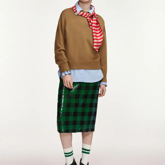 A model stands against a light gray studio background wearing a brown sweater, a red and white striped neck scarf, a light blue striped button-up shirt, a green sequined plaid midi skirt, green-and-white striped socks, and black patent leather high-heeled boots.