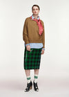 A model stands against a light gray studio background wearing a brown sweater, a red and white striped neck scarf, a light blue striped button-up shirt, a green sequined plaid midi skirt, green-and-white striped socks, and black patent leather high-heeled boots.
