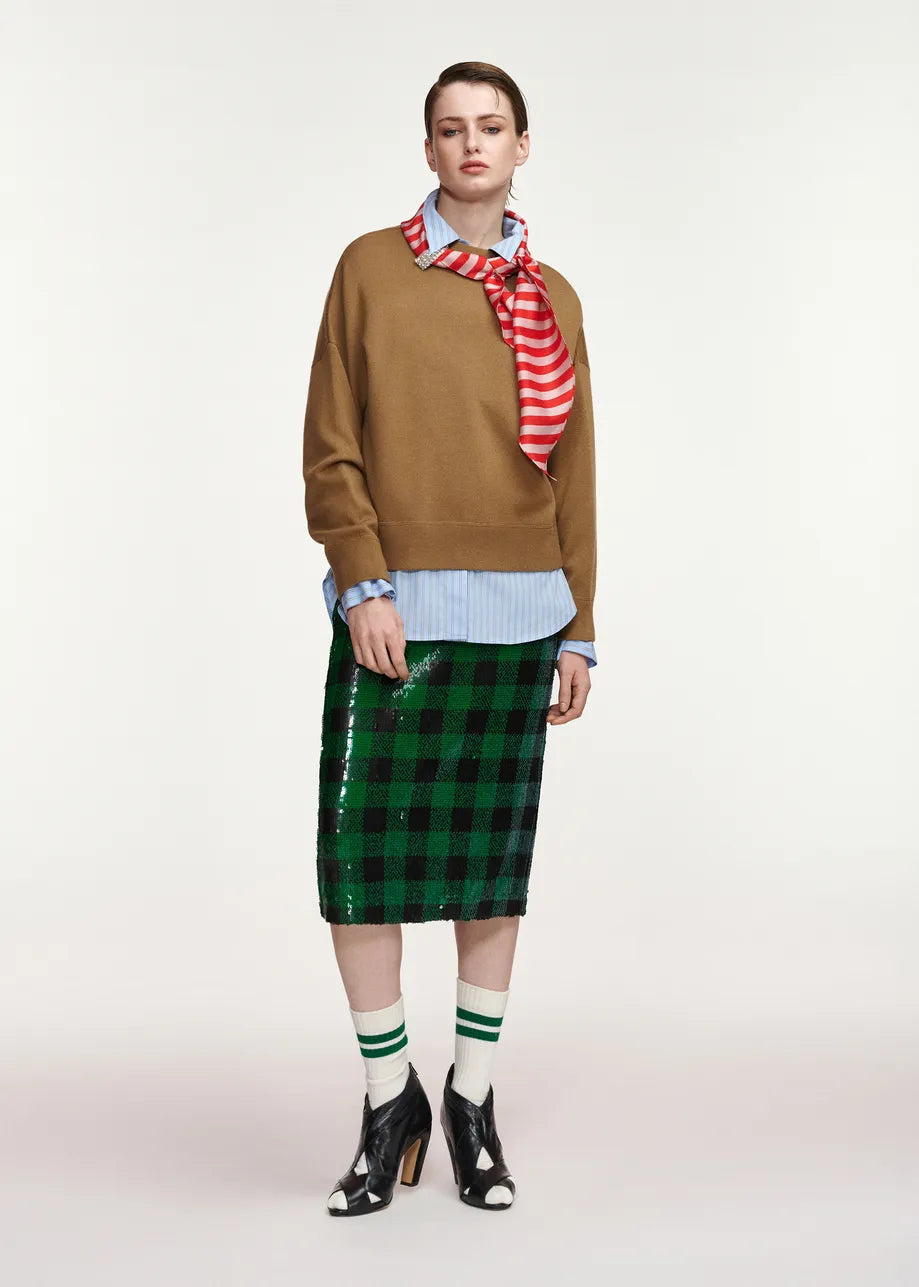 A model stands against a light gray studio background wearing a brown sweater, a red and white striped neck scarf, a light blue striped button-up shirt, a green sequined plaid midi skirt, green-and-white striped socks, and black patent leather high-heeled boots.