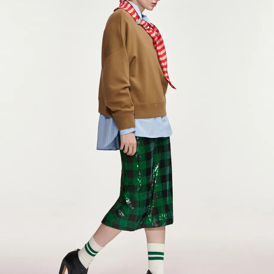A model is shown in profile wearing a brown cropped sweatshirt, a red and white striped neck scarf, a light blue button-down shirt, a green sequined plaid midi skirt, white socks with green stripes, and black high-heeled shoes, against a plain light gray studio background.