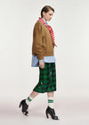 A model is shown in profile wearing a brown cropped sweatshirt, a red and white striped neck scarf, a light blue button-down shirt, a green sequined plaid midi skirt, white socks with green stripes, and black high-heeled shoes, against a plain light gray studio background.