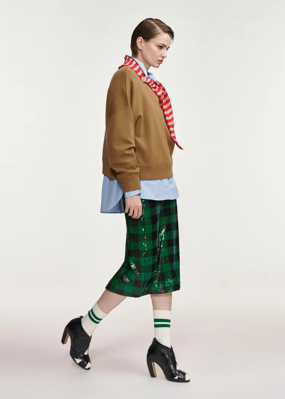 A model is shown in profile wearing a brown cropped sweatshirt, a red and white striped neck scarf, a light blue button-down shirt, a green sequined plaid midi skirt, white socks with green stripes, and black high-heeled shoes, against a plain light gray studio background.