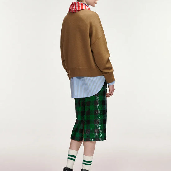 A model is shown from the back wearing a brown knit sweater, a light blue striped shirt, a red and white checkered neck warmer, a green sequined plaid midi skirt, green and white striped socks, and black high-heeled boots, against a plain light gray studio background.