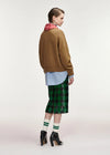 A model is shown from the back wearing a brown knit sweater, a light blue striped shirt, a red and white checkered neck warmer, a green sequined plaid midi skirt, green and white striped socks, and black high-heeled boots, against a plain light gray studio background.