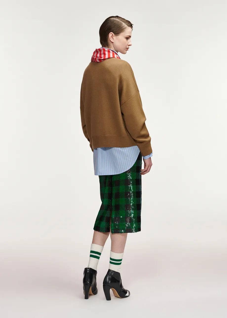 A model is shown from the back wearing a brown knit sweater, a light blue striped shirt, a red and white checkered neck warmer, a green sequined plaid midi skirt, green and white striped socks, and black high-heeled boots, against a plain light gray studio background.