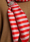 A close-up of a red and white striped silk scarf tied in a knot with a rhinestone-embellished band, resting against a brown textured fabric background.