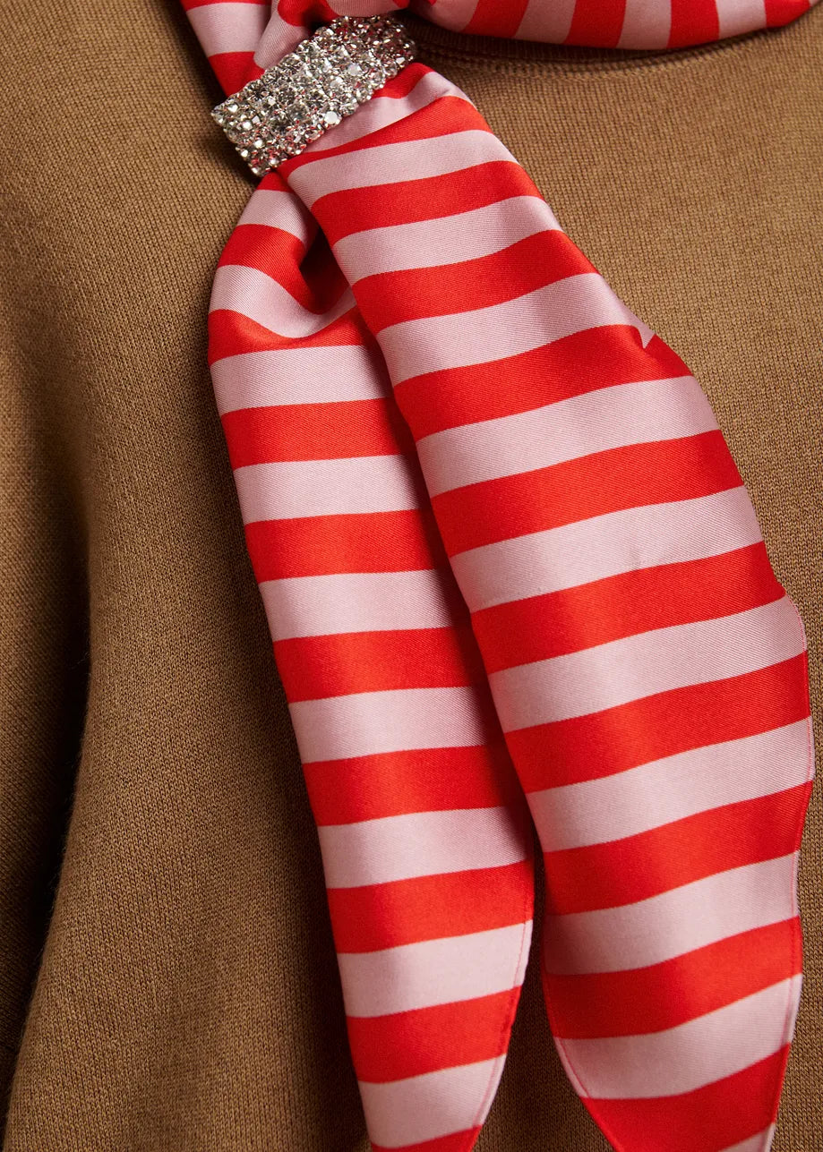 A close-up of a red and white striped silk scarf tied in a knot with a rhinestone-embellished band, resting against a brown textured fabric background.