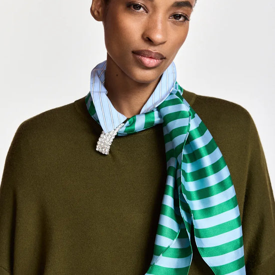 A model wearing a green and light blue striped silk scarf tied around a light blue striped shirt collar, paired with an olive green knit sweater, against a plain light gray background.