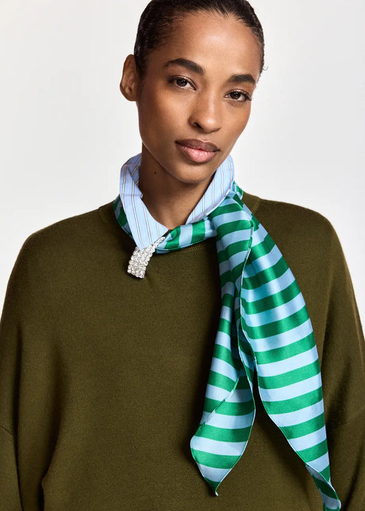 A model wearing a green and light blue striped silk scarf tied around a light blue striped shirt collar, paired with an olive green knit sweater, against a plain light gray background.