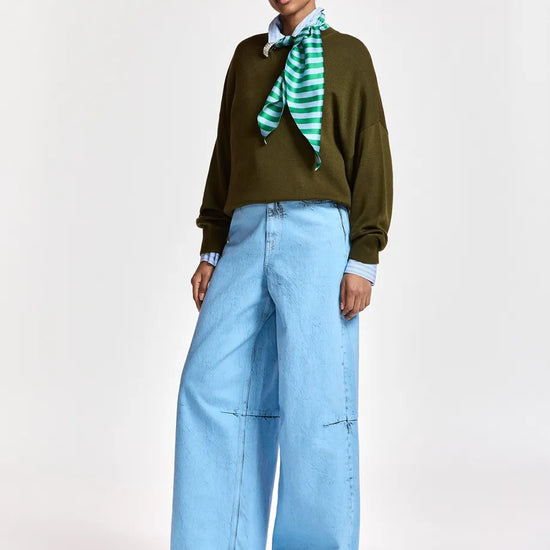 A model stands against a light gray studio background wearing a dark olive green long-sleeve top, light blue wide-leg jeans, a light blue collared shirt, a green and white striped neck scarf, and black shoes.