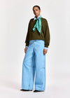 A model stands against a light gray studio background wearing a dark olive green long-sleeve top, light blue wide-leg jeans, a light blue collared shirt, a green and white striped neck scarf, and black shoes.