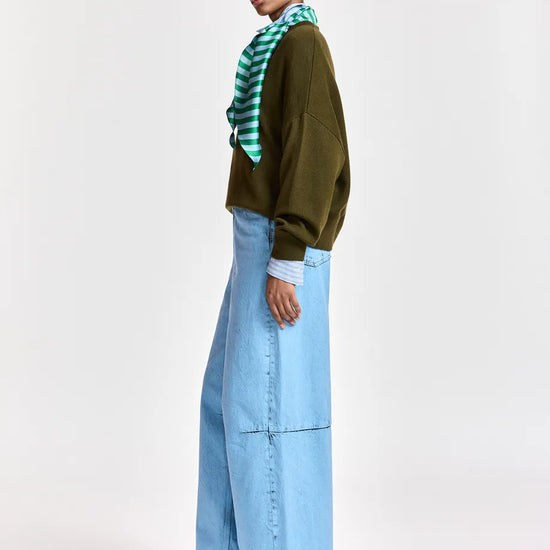 A model is shown from the side wearing a dark olive green long-sleeve top, a green and white striped neck scarf, and light blue wide-leg denim jeans against a plain light gray studio background.