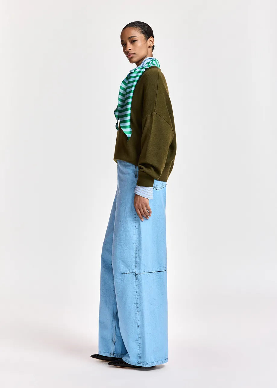 A model is shown from the side wearing a dark olive green long-sleeve top, a green and white striped neck scarf, and light blue wide-leg denim jeans against a plain light gray studio background.