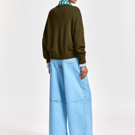 A model is shown from the back wearing an olive green crewneck sweater, wide-leg light blue denim pants, and black pointed shoes, set against a plain white studio background.