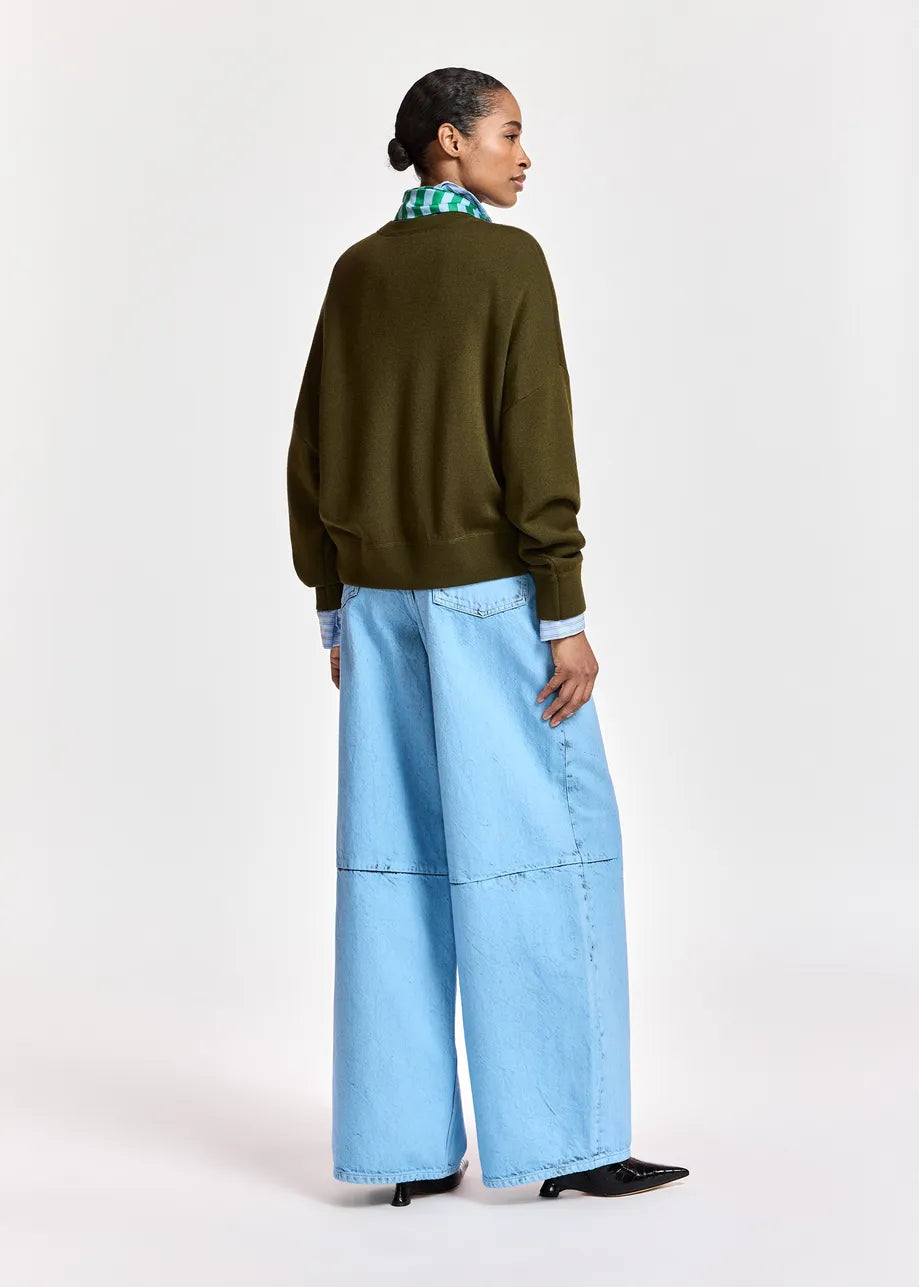 A model is shown from the back wearing an olive green crewneck sweater, wide-leg light blue denim pants, and black pointed shoes, set against a plain white studio background.