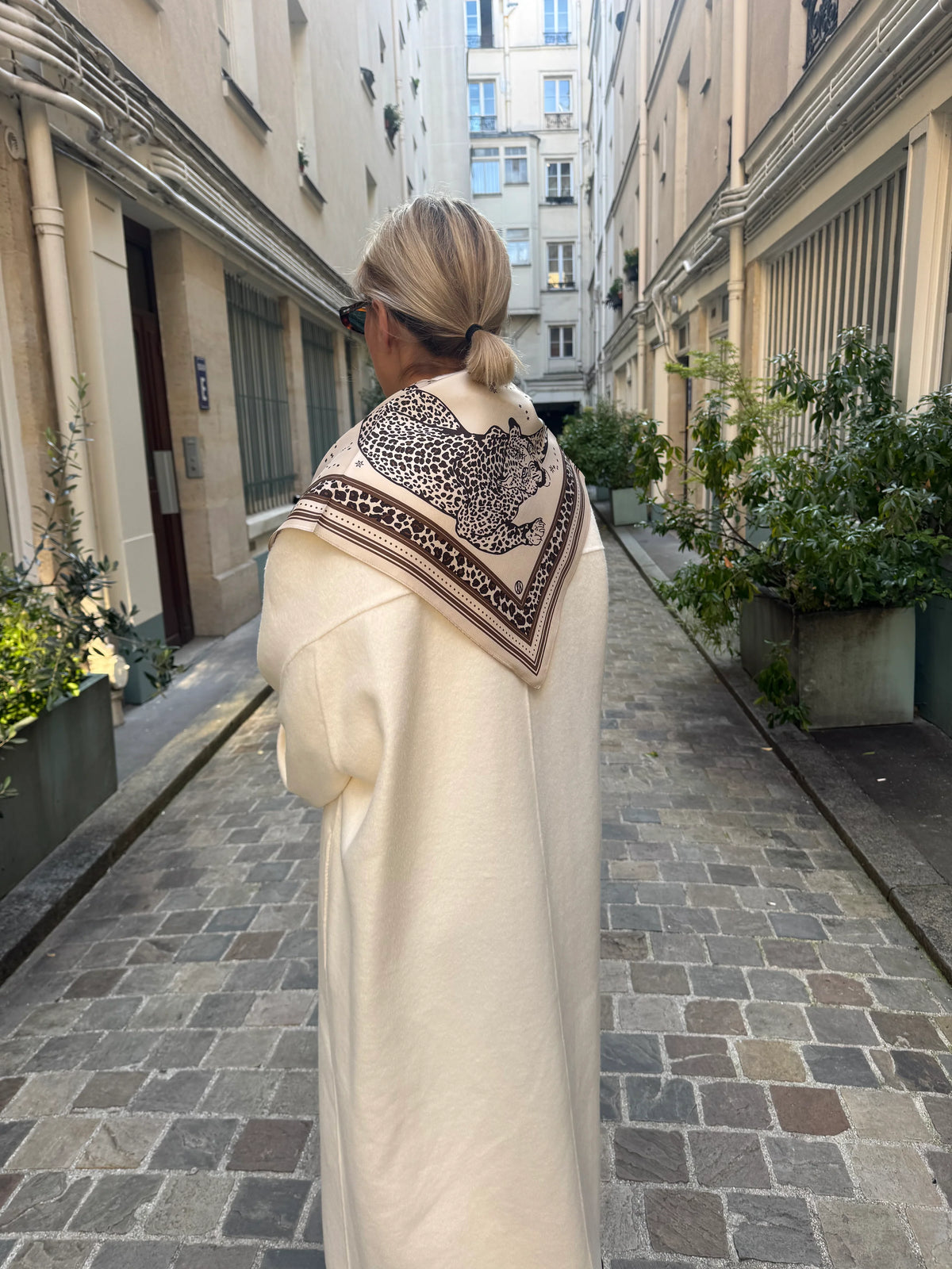 Woman walking down a narrow street wearing a long coat and patterned scarf.
