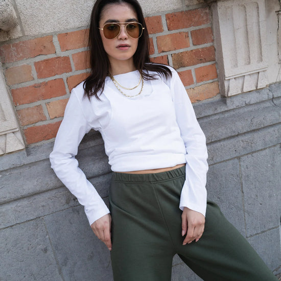 Woman wearing a white long-sleeve top and green pants against a brick wall.