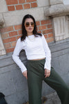 Woman wearing a white long-sleeve top and green pants against a brick wall.