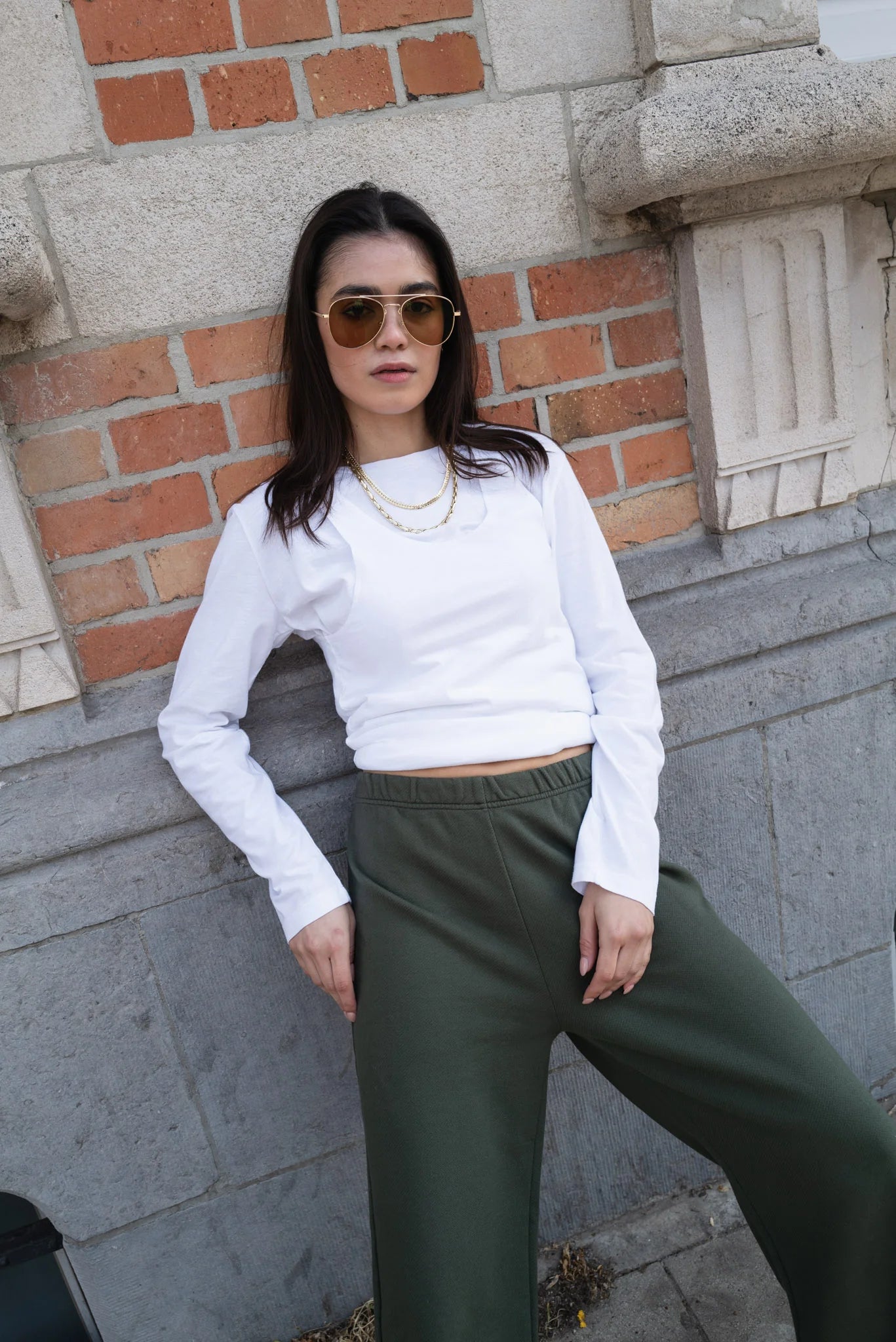 Woman wearing a white long-sleeve top and green pants against a brick wall.