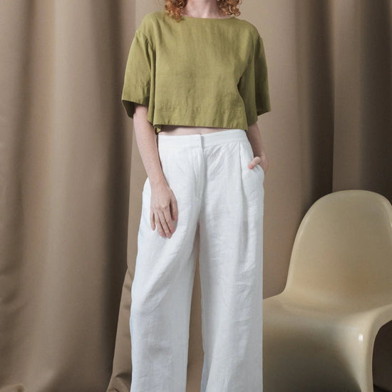 Woman wearing a green top and white pants standing against a beige curtain with a chair.