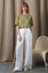 Woman wearing a green top and white pants standing against a beige curtain with a chair.