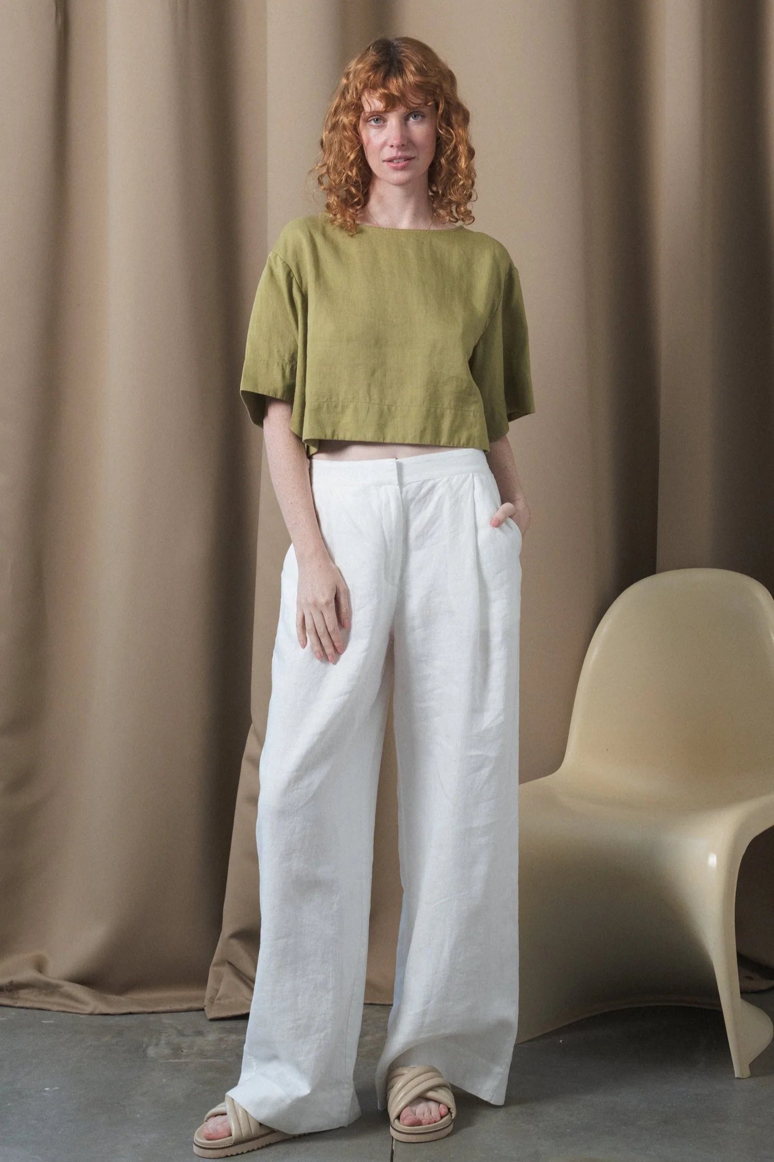 Woman wearing a green top and white pants standing against a beige curtain with a chair.