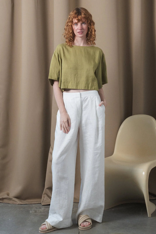 Woman wearing a green top and white pants standing against a beige curtain with a chair.
