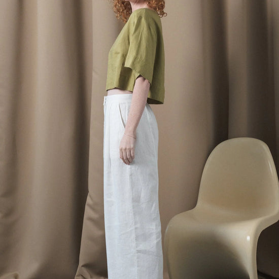 Woman wearing a green top and white pants standing against a beige curtain with a beige chair.