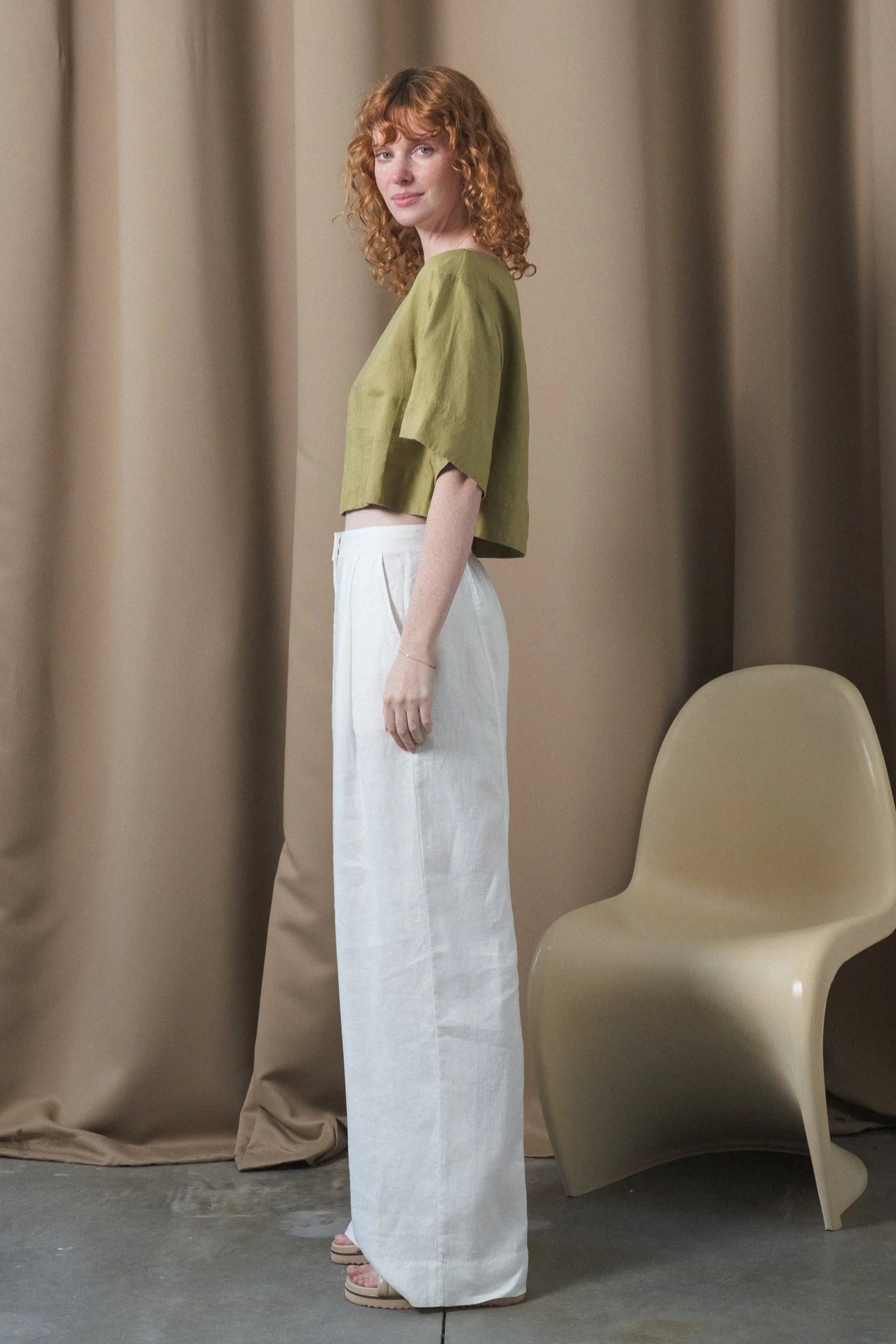 Woman wearing a green top and white pants standing against a beige curtain with a beige chair.
