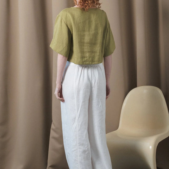 Woman wearing a green top and white pants standing in front of a beige curtain with a white chair.