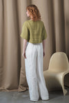 Woman wearing a green top and white pants standing in front of a beige curtain with a white chair.