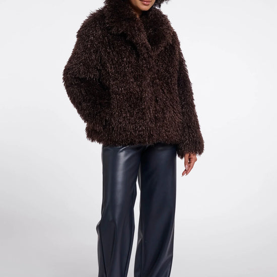 A model wearing a dark brown shaggy faux-fur jacket, dark navy wide-leg faux-leather pants, and beige pointed-toe flats, standing against a light gray studio background.